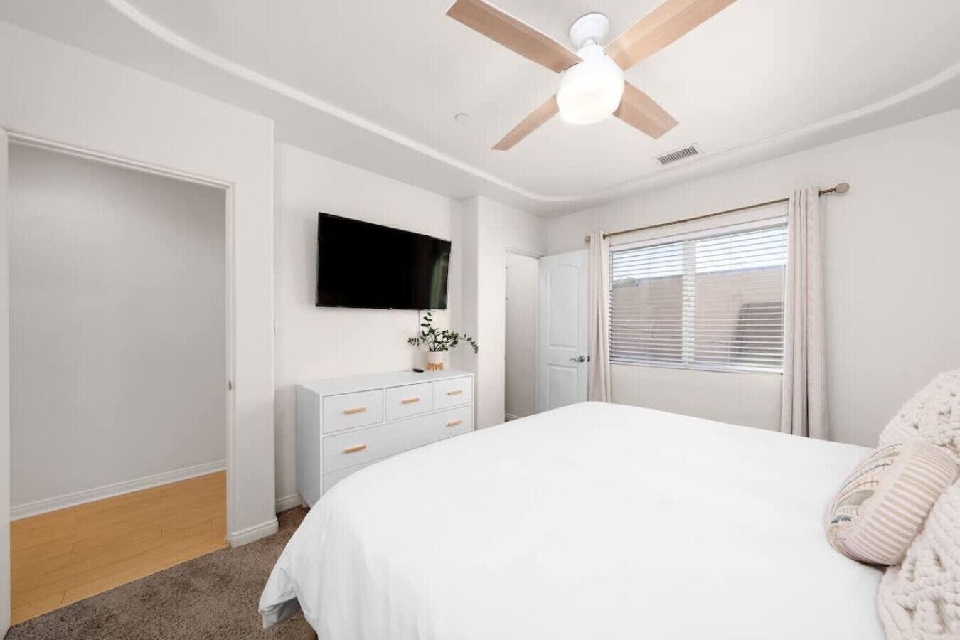 Un dormitorio con cama, cómoda, televisor, ventilador de techo y ventana con persianas.