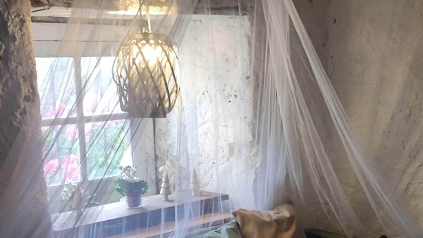 A room with a rustic stone wall, a wooden bench with cushions, a hanging light fixture, and a window with a view of greenery.