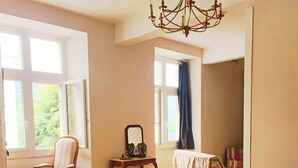 A dining area with a round table, chairs, a chandelier, and a view of greenery outside.