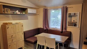 Une pièce de taille modeste, comprenant un meuble en bois, un banc, une table avec des chaises, une fenêtre avec des rideaux et un climatiseur fixé au mur.