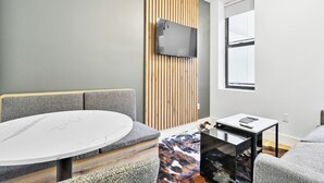 A modern room with a round table, grey sofa, wooden slat wall panel, mounted TV, and a window. - 1 Bedroom New Renovated - Wash/Dryer & Kitchenette -E (New York)