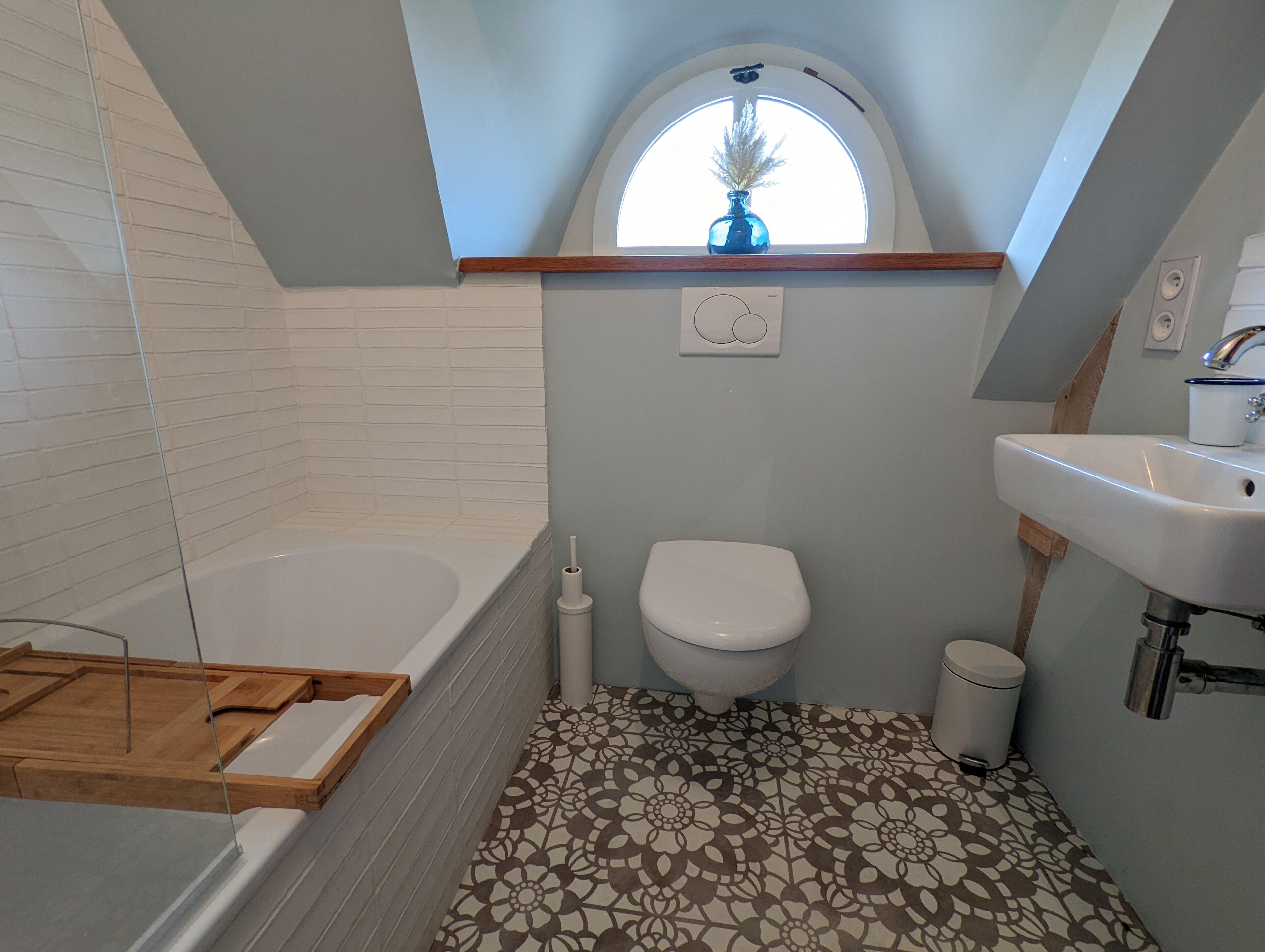 A bathroom with a bathtub, toilet, and sink, featuring a patterned floor and a small window with a vase.