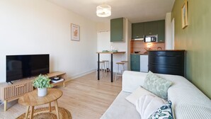 A modern living room with a flat-screen TV, a sofa, a coffee table, and a kitchen area with green cabinets and a microwave. - Le Soleil Bleu - à 50m de la plage (Courseulles-sur-Mer)
