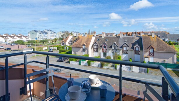 A balcony with a table set for two, overlooking a street with traditional houses and a clear sky. - Le Soleil Bleu - à 50m de la plage (Courseulles-sur-Mer)