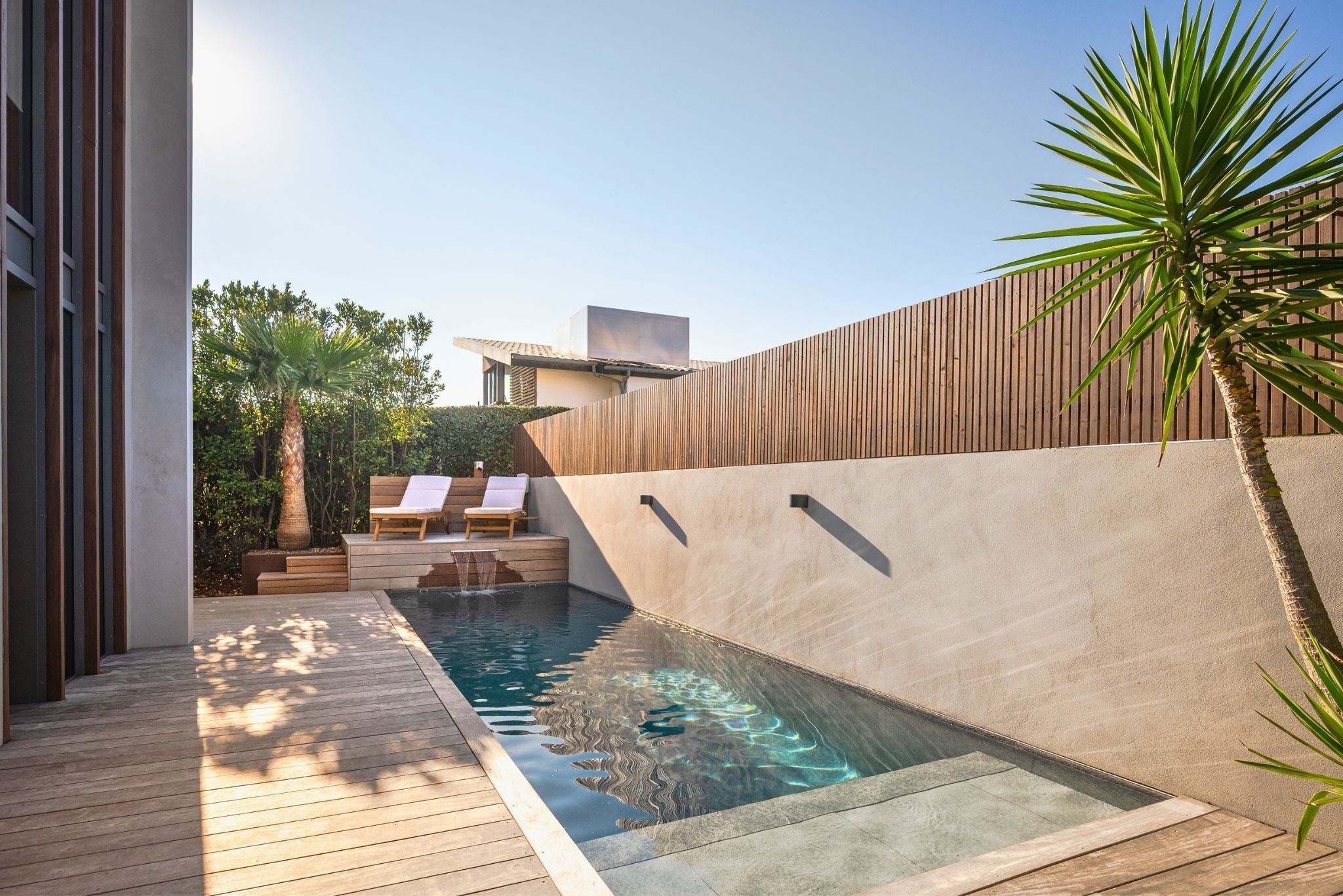Un área de piscina moderna al aire libre con cubierta de madera, sillas de descanso y un muro de madera.