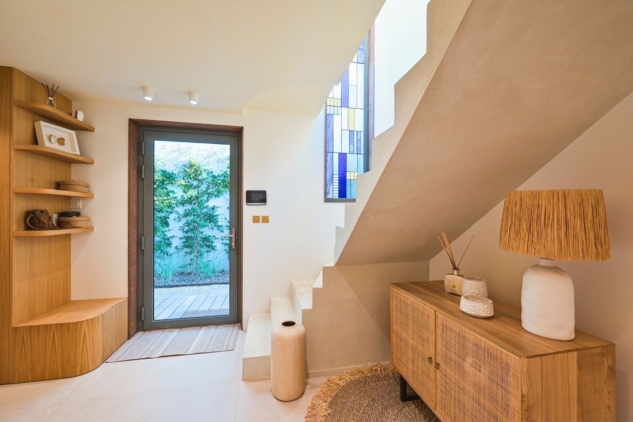 Un interior moderno con una escalera de madera, un aparador con objetos decorativos y un ventanal con vistas al verdor.