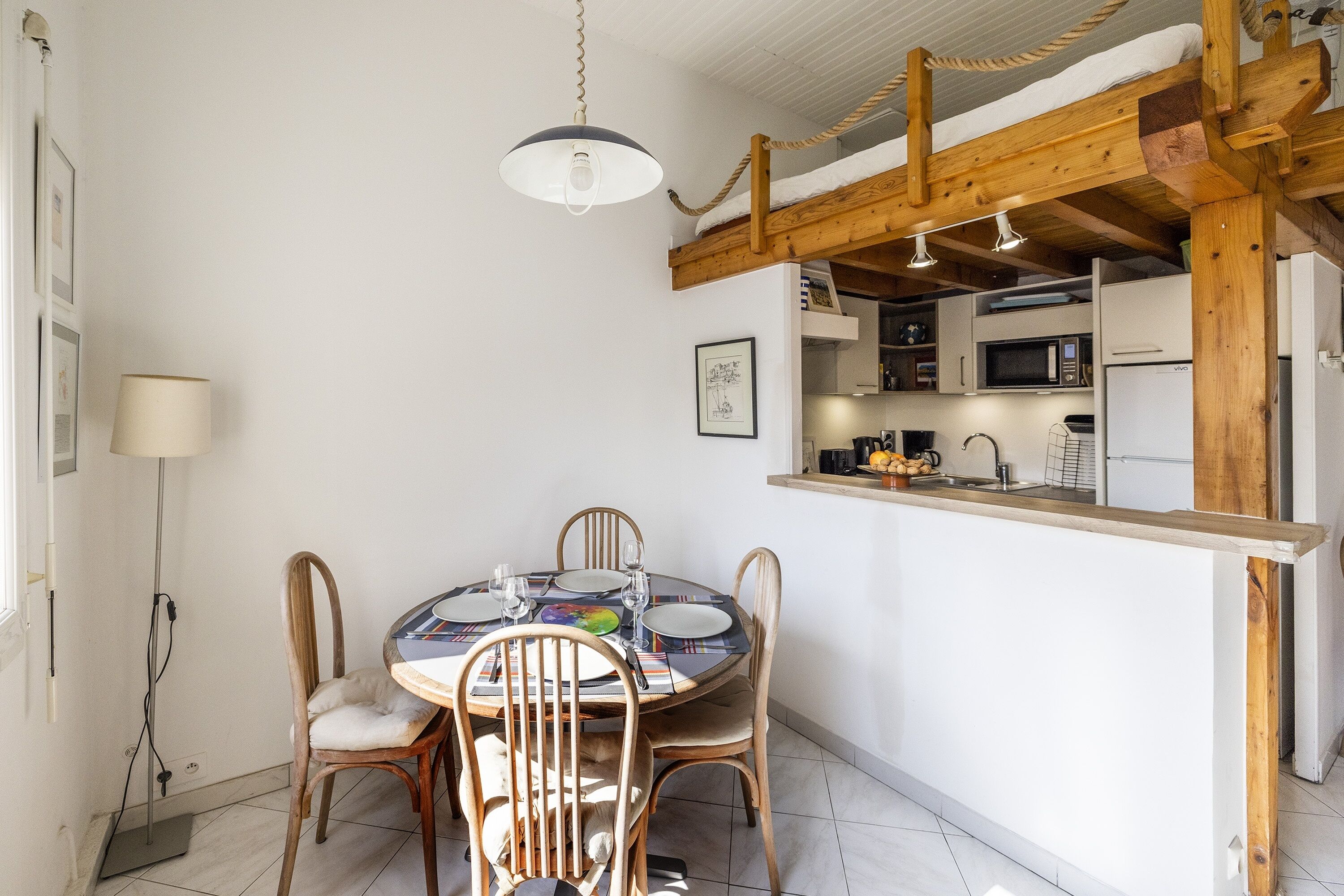 A dining area with a round table, four chairs, a hanging light fixture, and a kitchen area with a microwave and refrigerator.
