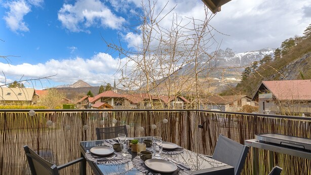 An outdoor dining area with a table set for four, surrounded by a bamboo fence, overlooking a mountainous landscape with houses in the background. - Le Cocon Élégance - à 500m du lac (Duingt)