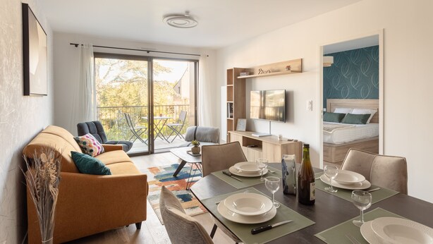 Ein modernes Wohnzimmer mit einer Couch, einem Esstisch für zwei Personen, einem Fernseher und einem Balkon mit Blick.
