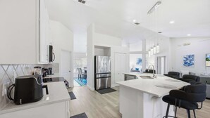 Una cocina moderna con armarios blancos, una isla central con taburetes y un refrigerador empotrado, además de un área de comedor visible a través de un vano abierto.