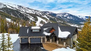 A house with a garage, stone foundation, and a view of snow-covered mountains. - Lodge with Incomparable Views & Direct Ski Access (Whitefish)