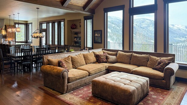 A spacious living room with a large sectional sofa, a dining area with a wooden table and chairs, and large windows offering mountain views. - Lodge with Incomparable Views & Direct Ski Access (Whitefish)