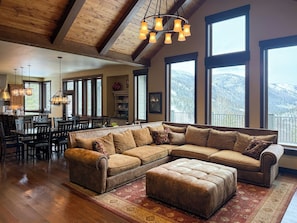 Un salon spacieux avec un grand canapé d’angle, un coin repas comprenant une table et des chaises en bois, et de grandes fenêtres offrant une vue sur les montagnes.