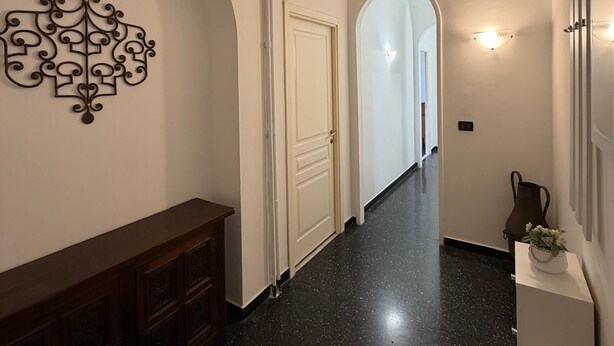 Un pasillo con piso de aspecto moteado y oscuro, paredes blancas, un adorno en la pared, una puerta y una mesita con un jarrón y zapatillas.