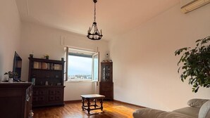 Un salón amplio con suelo de madera, un ventanal grande, una araña, un sofá y una planta.