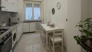 Una cocina compacta con microondas, estufa y pileta, una mesa de comedor con sillas, un ventanal con cortinas y una planta en maceta.