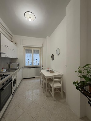 Eine kleine Küche mit Mikrowelle, Herd und Spüle, ein Esstisch mit Stühlen, ein Fenster mit Vorhängen und ein Blumentopf.