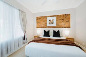 A bedroom with a large bed, a brick accent wall, a framed picture, two bedside lamps, and sheer curtains. - Percys Cottage Storybook Heritage Charm in Town (Mudgee)