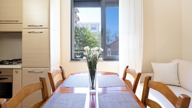 A dining area with a table, chairs, a vase with flowers, a window with a view of buildings, and a kitchen area. - Sea Point Apartments (Durrës)