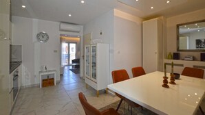 Un comedor moderno con una mesa blanca, sillas anaranjadas, un reloj y una cocina al fondo.