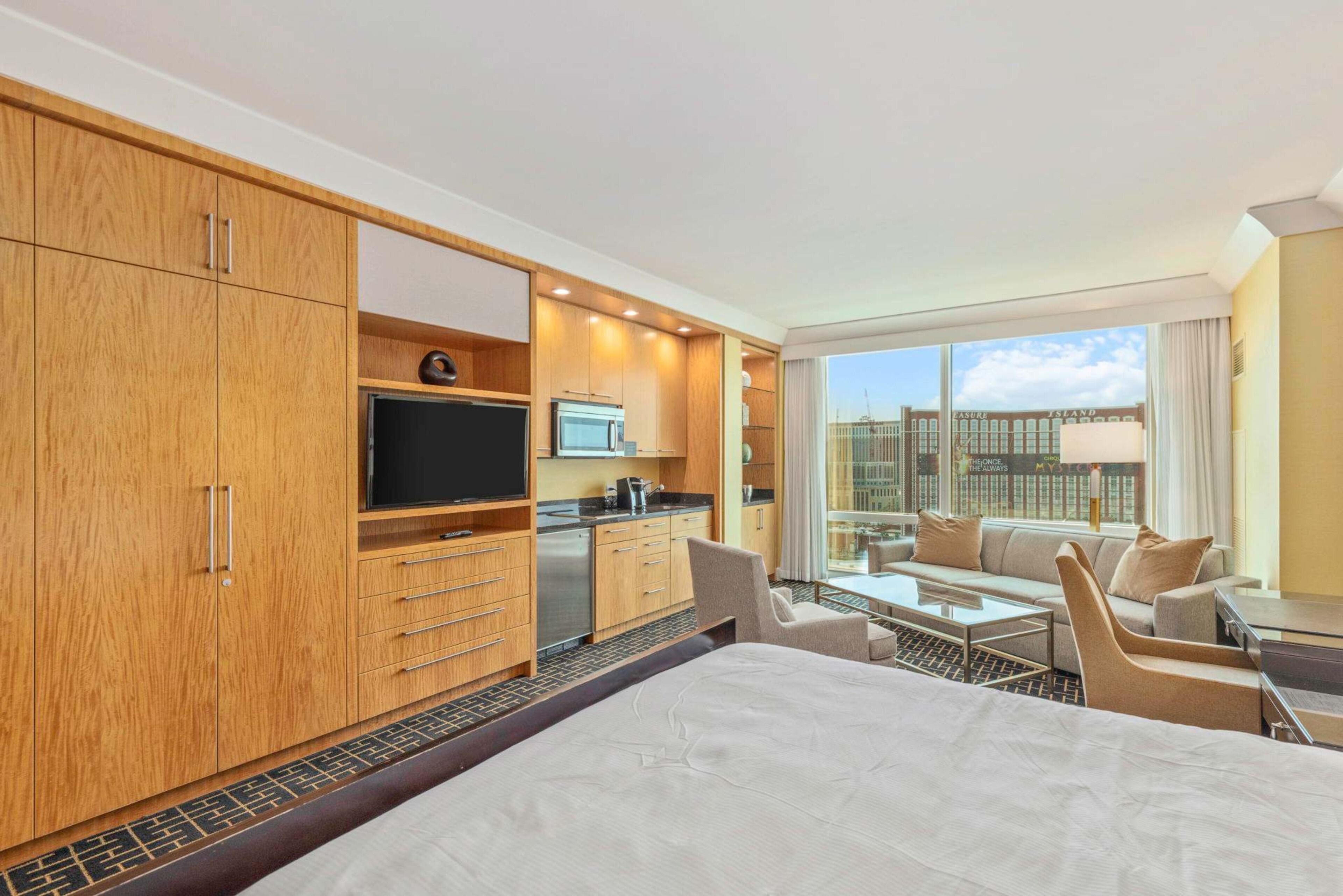 A hotel room with a large bed, a kitchenette, a TV, and a city view.