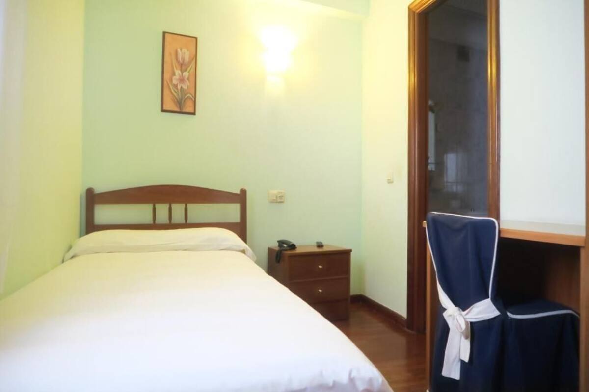 A room with a bed, a nightstand, a chair, a wall-mounted light, and a door.