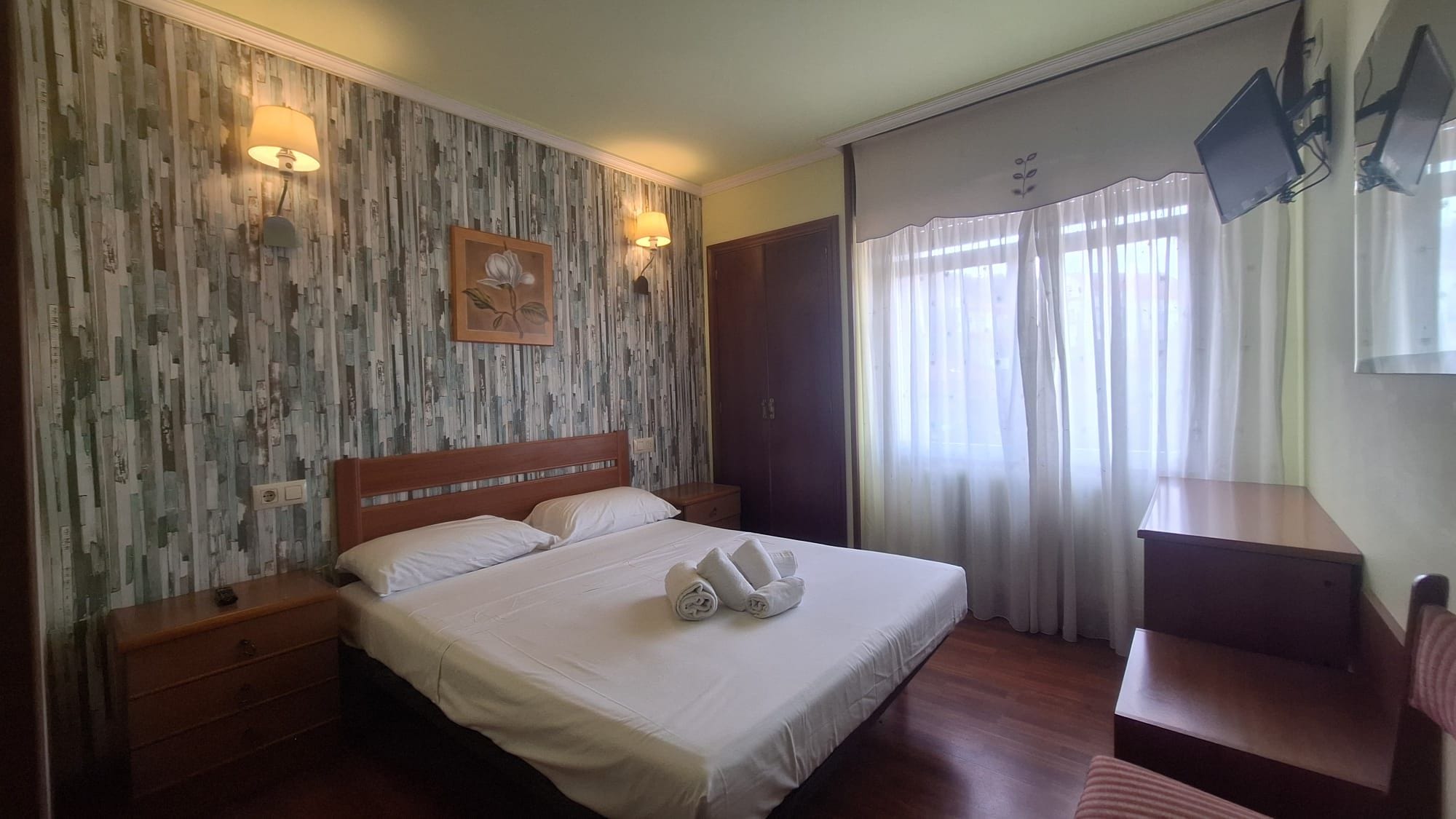Une chambre d’hôtel avec un lit, des lampes de chevet, une table de chevet en bois, un bureau avec une chaise et une grande fenêtre avec des rideaux.