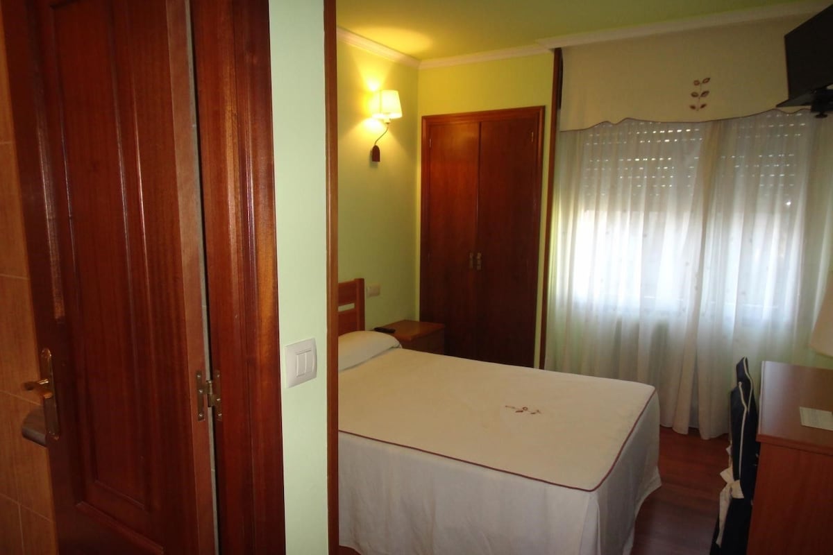 Una habitación de hotel con cama, mesita de noche, armario, lámpara y ventana con cortinas.