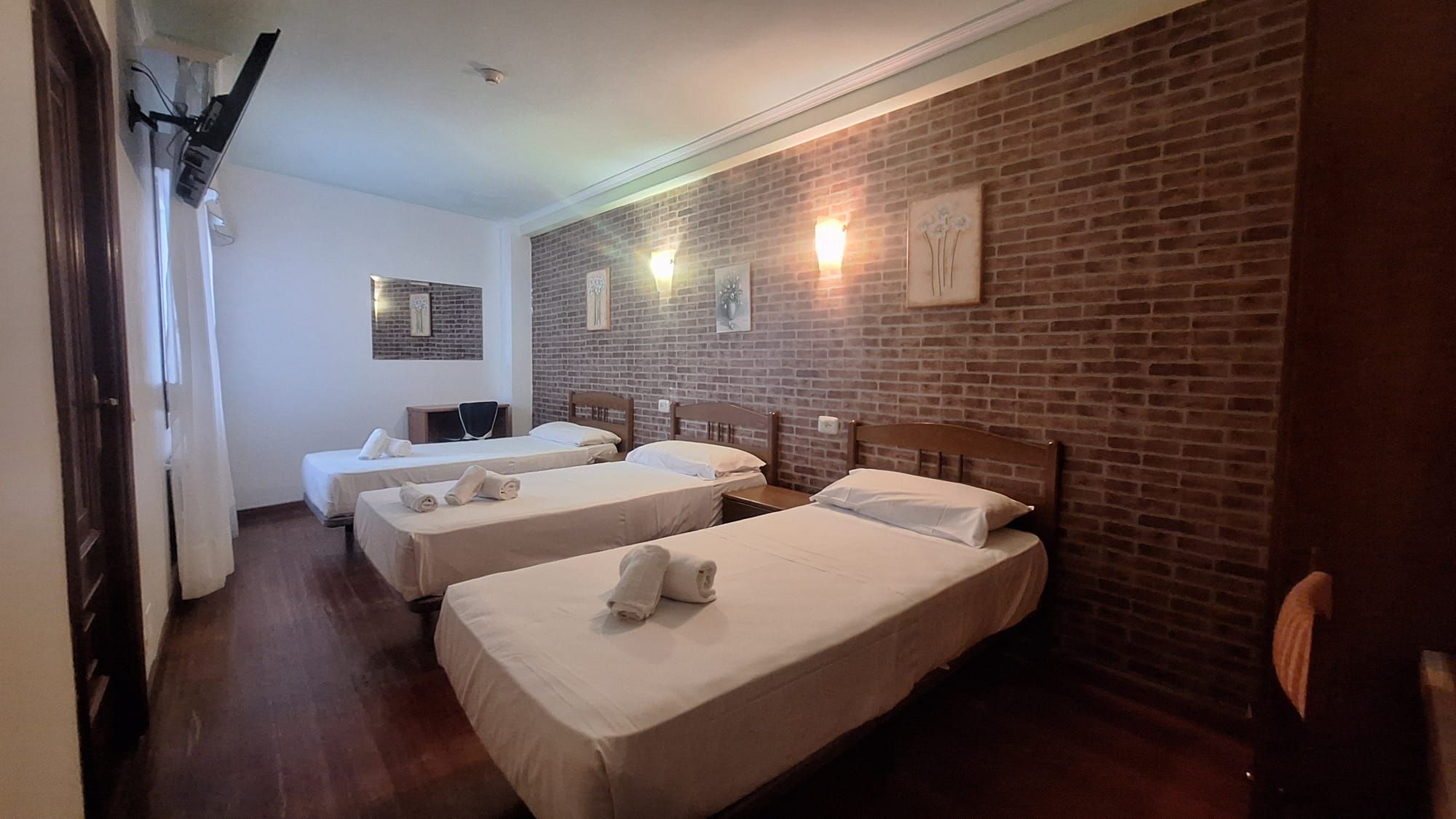 Habitación de hotel con tres camas individuales, una pared de ladrillo y un televisor de pantalla plana montado en la pared.