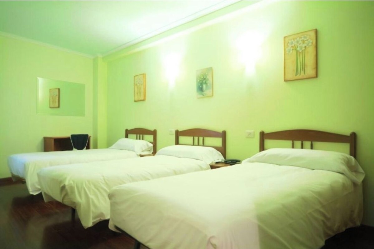 Una habitación con tres camas individuales, cada una con cabecera de madera, ropa de cama blanca y una mesita de noche pequeña.