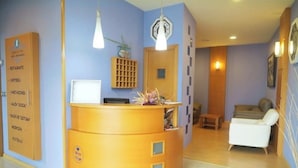A reception desk with a computer, a clock, and a plant. There is a white chair and a wooden cabinet with a grid pattern. - My Rooms Hotel Camiño do Mar (Rois)