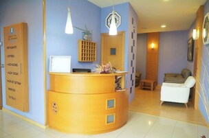 Mostrador de recepción con una computadora, un reloj y una planta. Hay una silla blanca y un mueble de madera con diseño de rejilla.