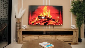 Een woonkamer met een televisie waarop een haard te zien is, een houten tv-meubel, een ronde salontafel met een boek en een potplant.