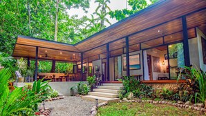 Ein modernes Haus mit überdachter Terrasse, umgeben von üppiger Vegetation und einem Kiesweg zum Eingang.