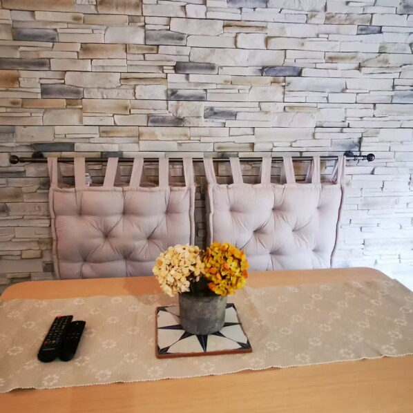 A room with a stone wall, tufted headboard, a table with a vase of flowers, and a remote control. - Mini house with full kitchen (Campan)