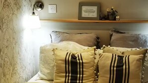 A bed with a checkered bedspread, multiple pillows, a bedside lamp, and a shelf with decorative items. - Mini house with full kitchen (Campan)