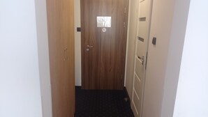 Un pasillo estrecho con una puerta de madera al final, alfombra negra y pared blanca.