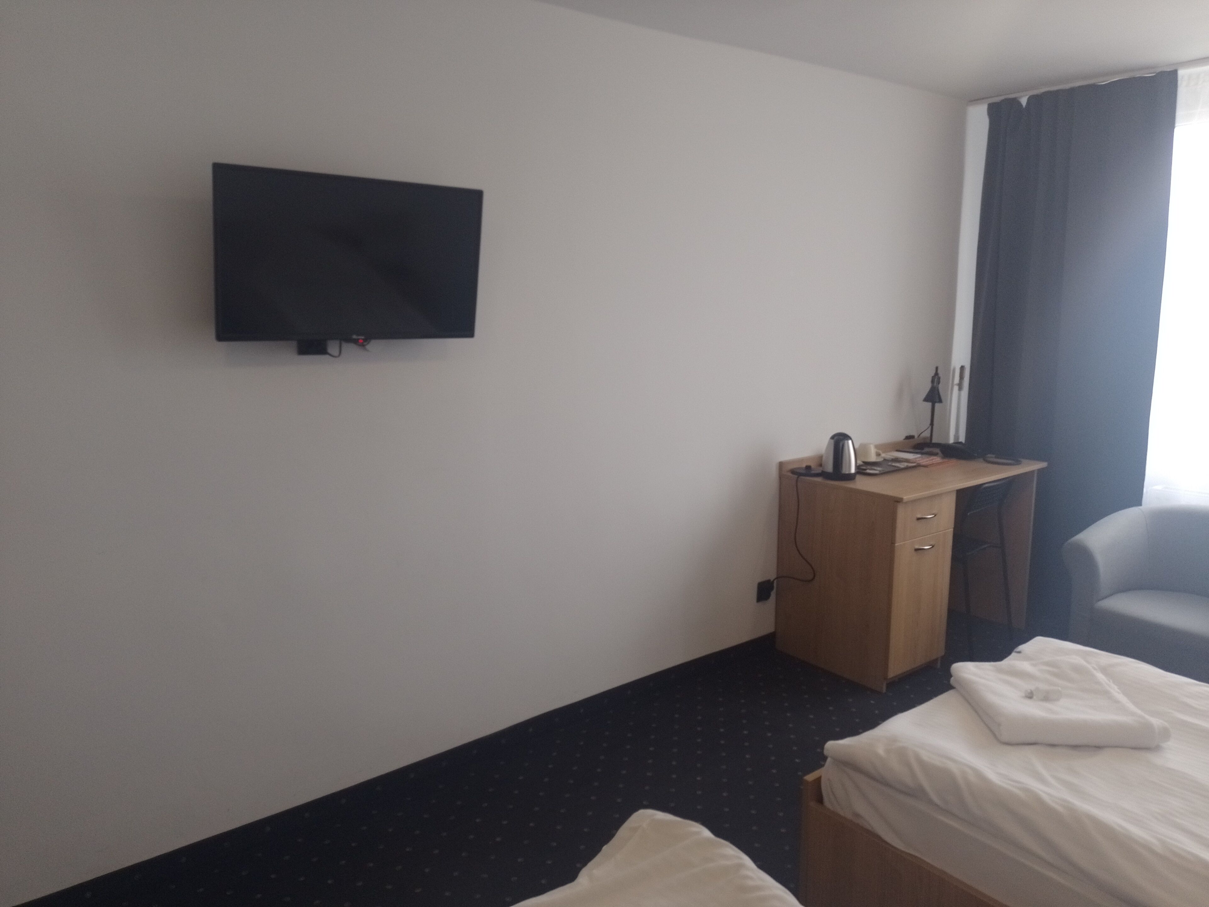 A hotel room with a flat-screen TV mounted on the wall, a wooden desk with a kettle and a cup, a chair, and two beds with white linens.