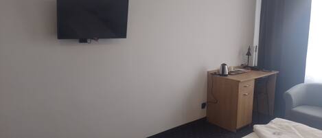 Habitación de hotel con televisión de pantalla plana montada en la pared, un escritorio de madera con hervidor y una taza, una silla y dos camas con ropa blanca.