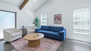 A modern living room with a blue sofa, a white armchair, a round coffee table, and a large window with blinds. - Oaks Cottages Modern 2BR Retreat (Lafayette)