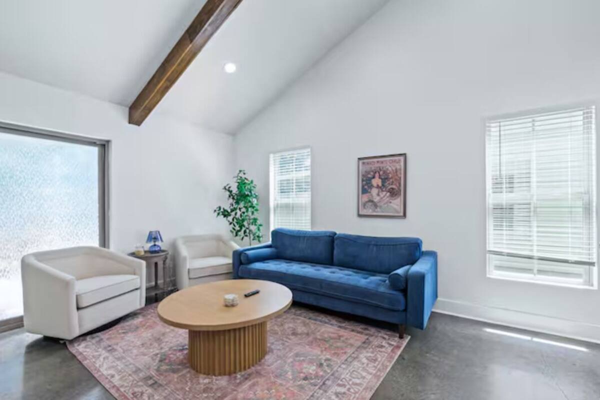 Een moderne woonkamer met een blauwe bank, een witte fauteuil, een ronde salontafel en een groot raam met zonwering.