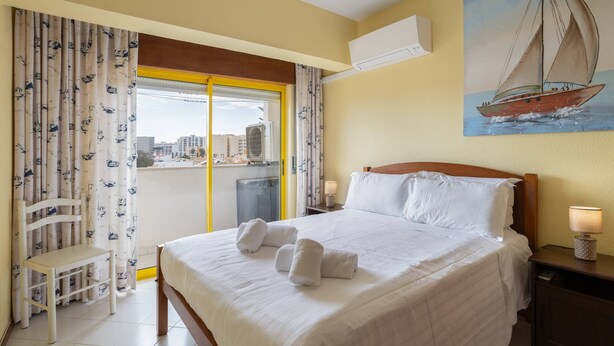 A hotel room with a bed, two bedside tables with lamps, a chair, a window with curtains, and a painting of a sailboat. - House Feliz by Centralgarve (Loulé)