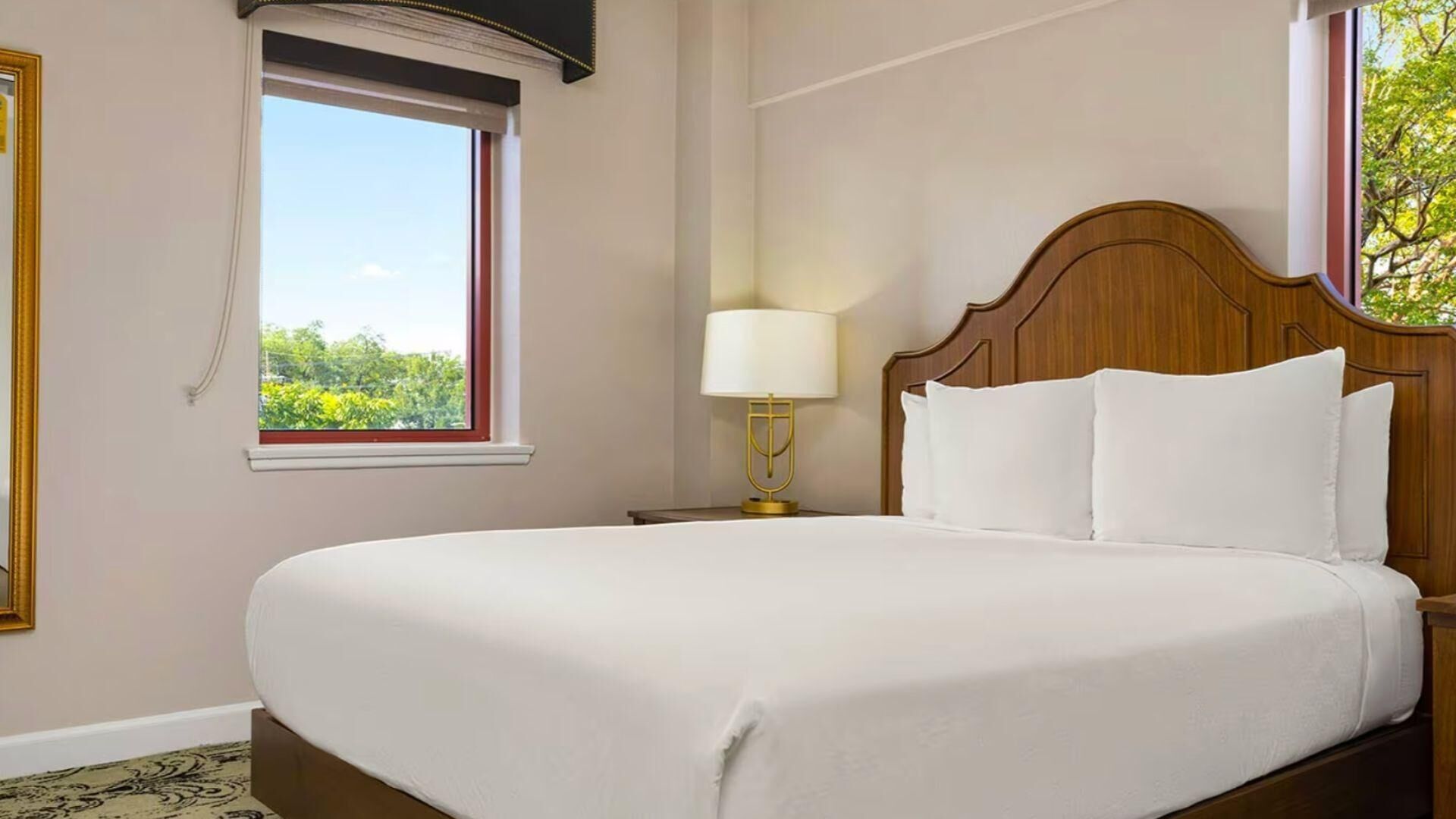A neatly made bed in a hotel room with a wooden headboard, a bedside lamp, and a window with a view of greenery.