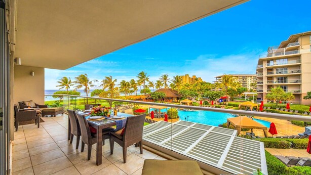 A balcony with a dining table set for two, overlooking a pool and a tropical landscape with palm trees and a view of the ocean. - Honua Kai 4 Free Rental Cars KBM Resorts Grand Suite 4 Units, 9 Bedrooms HK Ml-2663 (Kaanapali)