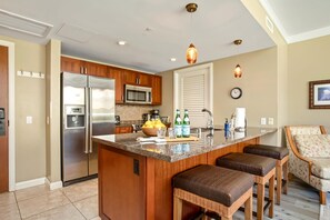 A kitchen with a central island, wooden cabinets, stainless steel appliances, and a dining area with wicker chairs. - Honua Kai Close To Beach 3 Free Rental Cars KBM Resorts 3 Units, 6 Bedrooms HK Ml-2666 (Kaanapali)