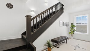 Una escalera con barandal de madera oscura y escalones, una pared blanca con el letrero 'Imported from Detroit', una planta en maceta, un banco y un suelo estampado.