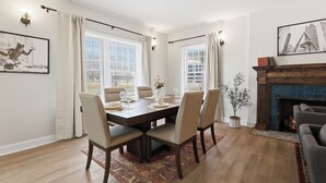 Comedor con una mesa de madera dispuesta para seis comensales, flanqueada por dos ventanas con cortinas, chimenea y vistas a la ciudad.