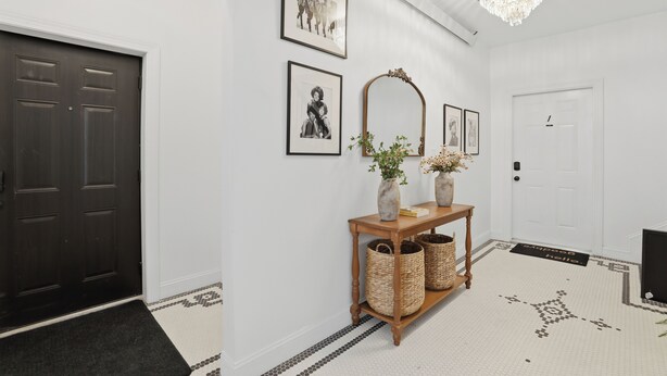 Un pasillo con una puerta oscura, una pared blanca con cuadros enmarcados, una consola de madera con jarrones y una alfombra estampada.
