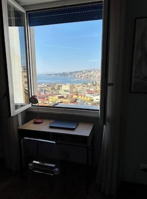 Ein Zimmer mit Schreibtisch, Stuhl und Blick auf eine Küstenstadt durch ein großes Fenster.