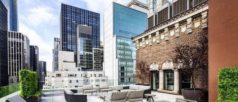 A rooftop terrace with outdoor seating, a brick building with classical architecture, and modern high-rise buildings in the background.